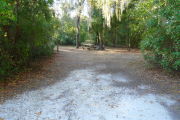 View of a vacant campsite with a picnic table, fire ring, and electric pedestal. The main part of the site is sandy with some small patches of grass. There are bushes on each side of the entrance and several medium size trees at the back of the site. There is partial shade.
