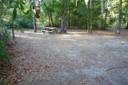 View of a vacant campsite with a picnic table, fire ring, and electric pedestal. The main part of the site is sandy with some small patches of grass. Several medium size trees are located at the back of the site. There is partial shade.
