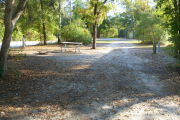 View of a vacant campsite with a picnic table, fire ring, electric pedestal, and water spigot. The main part of the site is sandy and hard packed in some areas. There are small trees and bushes on both sides of the site and there is partial shade.