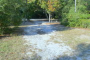 View of a vacant campsite with a picnic table, fire ring, electric pedestal, and water spigot. The main part of the site is sandy and hard packed in some areas. There are small trees and bushes on both sides of the site and there is partial shade.