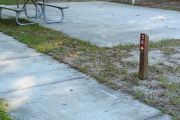 Close up of a brown site marker with an arrow on a wooden post. The site marker is to the right side of a sidewalk and a large concrete pad with a picnic table is present in the background.