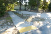 View of a vacant campsite with a picnic table, fire ring, electric pedestal, and water spigot. The site has a concrete side walk and camper pad. The sidewalk leads to the campsite and to the campground restroom. There are a few trees on the edges and partial shade. This site is ADA compliant.