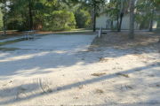 View of a vacant campsite with a picnic table, fire ring, electric pedestal, and water spigot. The site has a concrete side walk and camper pad. The sidewalk leads to the campsite and to the campground restroom. There are a few trees on the edges and partial shade. This site is ADA compliant.