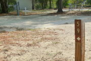 Close up of a brown site marker with an arrow on a wooden post. There is sand in the foreground and a concrete pad with a water spigot and electric pedestal are in the background.