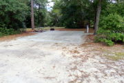 View of a vacant campsite with a picnic table, fire ring, electric pedestal, and water spigot. The site has a concrete side walk and camper pad. The sidewalk leads to the campsite and to the campground restroom. There are a few trees on the edges and partial shade. This site is ADA compliant.