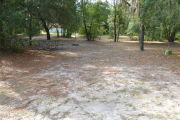 View of a vacant campsite with a picnic table, grill, and water spigot (this site does not have electricity).  The main part of the site is sandy with some patches of grass. There are trees spread throughout the site and there is partial shade.