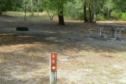 Close up of a brown site marker with an arrow on a wooden post. There is grass in the background, as well as, a picnic table and fire ring.