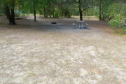 View of a vacant campsite with a picnic table, grill, and water spigot (this site does not have electricity).  The main part of the site is sandy with some patches of grass. There are trees spread throughout the site and there is partial shade.