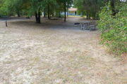 View of a vacant campsite with a picnic table, grill, and water spigot (this site does not have electricity).  The main part of the site is sandy with some patches of grass. There are trees spread throughout the site and there is partial shade. The campground restroom is present in the background.