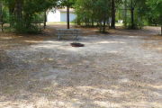 View of a vacant campsite with a picnic table, fire ring, and a water spigot (this site does not have electricity). The main part of the site is sand and there are a few trees around the site. There is partial shade and the campground restroom is present in the background.