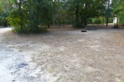 View of a vacant campsite with a picnic table, fire ring, and a water spigot (this site does not have electricity). The main part of the site is sand and there are a few trees around the site. There is partial shade and the campground restroom is present in the background.