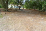 View of a vacant campsite with a picnic table, fire ring, and water spigot (this site does not have electricity). The main part of the site is sand and the edges of the site are lined with wooden barricades. There are trees scattered in the background and the campground restroom is behind the site.