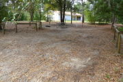 View of a vacant campsite with a picnic table, fire ring, and water spigot (this site does not have electricity). The main part of the site is sand and the edges of the site are lined with wooden barricades. There are trees scattered in the background and the campground restroom is behind the site.