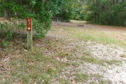 View of a vacant campsite with a brown site marker on a wooden post. There is grass in the foreground and a picnic table and fire ring are in the background.