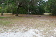 View of a vacant campsite with a picnic table, grill, and water spigot (this site does not have electricity).  The main part of the site is sandy with some patches of grass. There are trees spread throughout and there is partial shade. In the background there is a path leading out of the site.