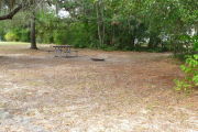 View of a vacant campsite with a picnic table and grill (this site does not have electricity).  The main part of the site is sandy with some patches of grass. There are trees spread throughout the site creating partial shade.