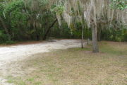 View of a vacant campsite with a picnic table, fire ring, electric pedestal, and water spigot. The main part of the site is sand with a large area of grass to the right side. There are two trees within the site and the back of the site is densely forested creating partial shade.