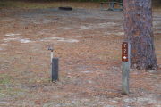 Close up of a brown site marker with an arrow on a wooden post. There is a water spigot to the left of the marker and a large tree directly behind it. A picnic table and fire ring is present in the background.