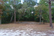 View of a vacant campsite with a picnic table, fire ring, water spigot, and electric pedestal. The main part of the site is sandy with patches of grass. There are several trees spread across the site and the back is densely forested creating partial shade.