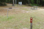 Close up of a brown site marker with an arrow on a wooden post. In the background a picnic table, water spigot, and fire ring are present. There are also two large grey electric boxes on the back edge of the site.