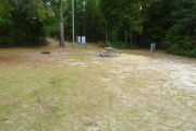 View of a vacant campsite with a picnic table, fire ring, and electric pedestal. The main part of the site is grass with some sandy areas mixed in. The side and back edges of the site are lined with trees creating partial shade. There are two large electric boxes at the back of the site.