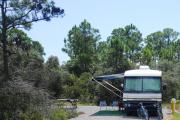One of the new 30/50 amp electric sites that has water and sewer. The site is gravel with a picnic and fire ring to the left and is surrounded with light vegetation.