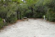 Front view of site 9 showing electric and water hook-up on the right side, clothesline to the left side, and picnic table and fire ring in the back. Site is surrounded by native vegetation. Site is dirt.