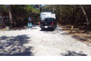Front view of site showing electric and water hook-up on the right.  Fire ring in the back of the site and picnic table in the middle. To the left is a clothesline. The site is surrounded by native vegetation. Site is dirt.