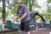 A man cooks breakfast on his small, green cookstove in front of his grey and yellow tent.