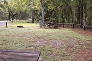 Partially sunny campsite with a fire ring, picnic table, and black top road.