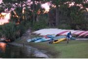 Photo: JONATHAN DICKINSON SP. The riverfront at Jonathan Dickinson State Park is shown in the golden sunset, as a man drags a kayak up the grassy bank in front of a line of beached canoes. His kayak has a bicycle strapped across the back of it. He is carrying a map.