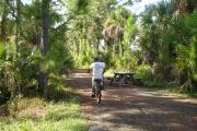 Photo: JONATHAN DICKINSON SP. Two people ride bicycles past a picnic table along a paved trail, under the shade of tall trees.