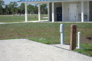 Photo: 023, Site number next to water and electric hooks ups at the back of a gravel site. A restroom is nearby.