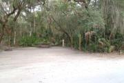 Lush vegetation of Spanish moss draped live oak, sable palms, and palmetto surrounding an open campsite with a cloths line, an electrical hook-up stanchion, a water spigot, a picnic table and a fire ring.