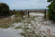 Photo: 001, Looking from the campground road toward the ocean, Small trees and other vegetation surrounds the campsite. The fire ring and fence rails are visible with the ocean behind them. White clouds drift across a blue sky.