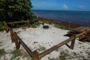 Photo: 002, View from shoreline toward campground road through the opening in the wooden fence rails. A picnic table and fire ring is shown on the sand and grass covered site. Green trees and plants surround the site in the buffer area between sites. A red tent is visible on the adjacent site.
