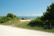 Photo: 003, View from road through campsite toward ocean. Fence rails a picnic table and campfire ring are visible with white puffy clouds in background over the green blue tinted ocean. A sign post on the right side has the site number and an arrow pointing to the site.