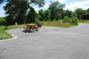 This is picture of campsite #61 from the back of the site looking toward the road.  This is an ADA accessible site that has 50amp electric, water and sewage hook-ups.  This is a sunny site with asphalt foundation and vegetation on sides.  There is a separate section to the side with the picnic table, cooking grill and ground campfire ring.  Next to the site is a sidewalk with easy access to the restroom. This site has to be pulled straight into.