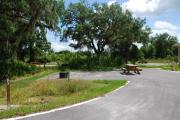 Campsite #63 is an ADA accessible, sunny site with asphalt pavement and vegetation on sides located on the right hand side of the road.  Looking into the site on the right are the water, 50 amp electric and sewage hook-ups. Also, to the right is a sidewalk to the restroom that is next to the site. To the left is a separate section containing an ADA accessible picnic table, cooking grill and ground campfire ring.  At the front left side of the site is the brown site marker.