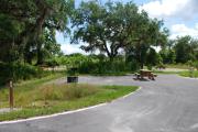 This picture of campsite #63 is looking from the road into the sunny site.  This is ADA accessible site has 50 amp electric, water and sewer hook-ups. There is an asphalt foundation with vegetation on the sides.  There is also a separate area to the side with a picnic table, cooking grill and ground campfire ring.  There is a sidewalk next to the site with easy access to the restroom.