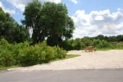 This picture of campsite #71 is looking from the road into the sunny site.  This site has 50 amp electric, water and sewer hook-ups. There is a stone foundation with vegetation on the sides.  There is also a separate area to the side with a picnic table, cooking grill and ground campfire ring.