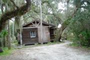 This is a front view picture of Cabin 1 which is a rustic palm log cabin built by the Civilian Conservation Corp. in the mid 1930âs.  This cabin is ADA accessible.  There is a living/dining/bedroom area with 2 double beds, a sofa bed, dining set, linen cabinet and fireplace.  There also is a bathroom and a kitchen with coffee maker, toaster, microwave, range, refrigerator, dishes, silverware and cooking utensils.  The cabins sleep up to six. Off of the back porch, there is a fire ring, cooking grill and a picnic table.  There are no pets and no smoking allowed in the cabins.