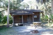 This is a back porch view picture of Cabin 4 which is a rustic palm log cabin built by the Civilian Conservation Corp. in the mid 1930âs.  There is a living/dining/bedroom area with 2 double beds, a sofa bed, dining set, linen cabinet and fireplace.  There also is a bathroom and a kitchen with coffee maker, toaster, microwave, range, refrigerator, dishes, silverware and cooking utensils.  The cabins sleep up to six.  Off of the back porch there is a fire ring, cooking grill and a picnic table.  There are no pets and no smoking allowed in the cabins.