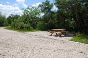 This is picture of campsite #86 from the back of the site looking toward the road.  Site has 50amp electric, water and sewage hook-ups.  This is a sunny site with stone foundation and vegetation on sides.  There is a separate section to the side with the picnic table, cooking grill and ground campfire ring.