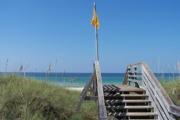 A boardwalk climbing through a dune lined by Sea Oats, leading to a beautiful white sandy beach where a yellow flag sways gently in the wind. The emerald green water glistens as it descends into a deep blue prism.
