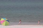 On a crystal clear day at St Andrews State Park, two young girls skip about on the white sandy beach with the emerald green water nipping at their toes. A colorful umbrella rest nearby.