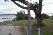 Sign post four situated to the right of an open campsite with a view of the lagoon in the distance. Electrical post and water spigot are located to the right of the post.