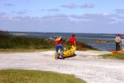 Photo shows youth camp boat ramp area and two young people carrying a kayak to the launch area.  Photo also shows other young people kayaking and others watching from the shore.