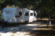 41x41 Foot site with water and electric hook ups. A white travel trailer with slide outs is parked on the shell rock pad shaded by large sprawling oaks that cover the entire site.