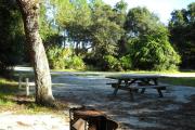 The view from the back of the site facing the road over looking fire grill and picnic table , oak tree on left side in front of water and electric hook ups.
